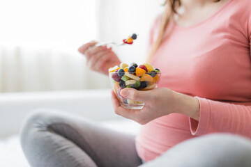 Pregnant female sitting in bedroom and eating fresh fruit salad.