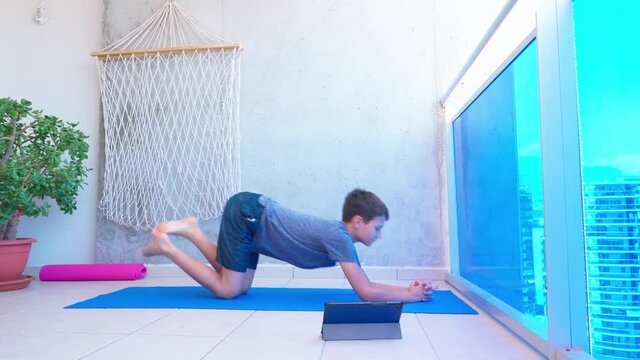 Kid doing sport exercises, doing plank exercise on yoga mat while watching video lesson online on tablet computer on balcony. Sport, healhty lifestyle, active leisure at home