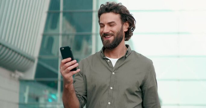 Attractive Handsome Man with nice Hairstyle and Beard Using his Modern Smartphone. Looking very stylish and Gorgeous. Wearing Khaki color Shirt and basic White Basic T-shirt. Typing Messages.