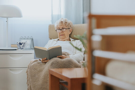 Senior woman sitting in armchair and reading book in nursing home