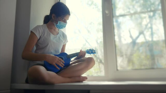 coronavirus pandemic. little teenage girl plays guitar ukulele sits on a windowsill by the window in a medical mask. self-isolation concept virus covid 19 infection doomsday. girl quarantined