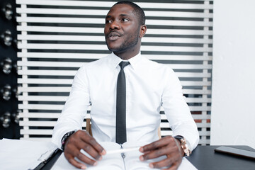 Fototapeta premium African American in a white shirt and black tie looks away while sitting at a table with documents, in the background a wooden panel