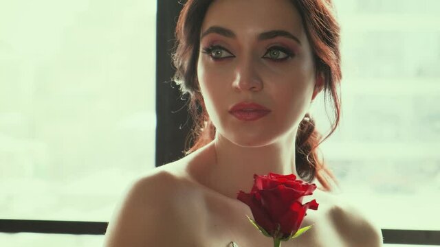 Closeup portrait young beautiful charming cute woman. Sexy bare shoulders. Evening makeup, pink lips, black eyebrows, beauty cats gray eyes, false eyelashes. Brunette girl holds red rose. loft window