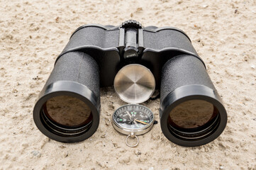 Black binoculars and a pocket compass on the sand.