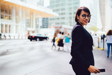 Half length portrait of happy businesswoman dressed in stylish formal wear and eyeglasses smiling at camera holding coffee to go standing on office building background near publicity area