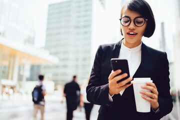 Smiling female manager surprised with getting discount of email holding blank coffee cup outdoors, prosperous businesswoman enjoying break sending messages on mobile standing on publicity area.