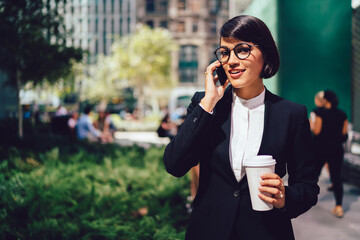 Half length portrait of positive woman economist in stylish eyewear and black suit communicating via cellphone during coffee break outdoors, cheerful female banker talking on smartphone in downtown