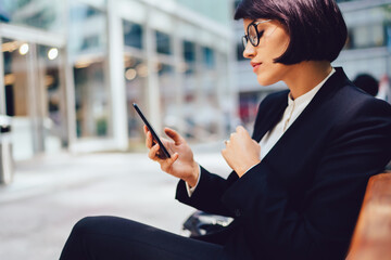 Side view of businesswoman in stylish outfit and spectacles resting on bench sending messages on smartphone.Confident owner of company using mobile application for banking online on cellular device