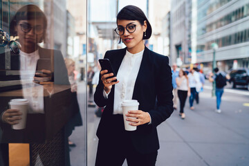 Half-length portrait of prosperous female owner walking on crowded street in downtown sending messages on smartphone, confident businesswoman holding coffee to go strolling looking at camera.