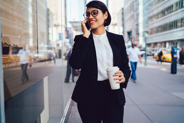 Positive young woman economist in stylish eyewear and black suite communicating via cellphone during coffee break outdoors, cheerful female banker talking with colleague via smartphone on street