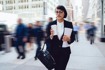 Pensive businesswoman walking around urban setting in financial district with coffee to go and report documents in hands, female banker in eyewear feeling pondering on way to office in New York city