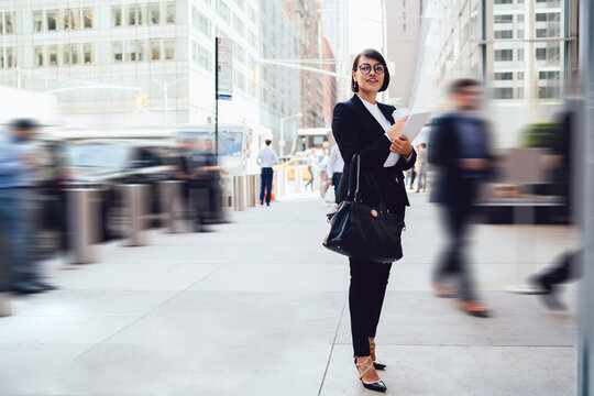 Positive woman in spectacles for vision correction standing on urban setting and enjoying work break before meeting with business partner, happy female economist in elegant suit looking away