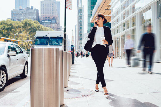 Confident woman in spectacles dressed in elegant suit strolling around modern streets in New York and using 4g internet connection for making international conversation with business partner