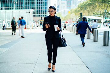 Young confident businesswoman in formalwear sending SMS on mobile while walking on city street, prosperous female manager reading news from networks on smartphone holding coffee to go cup strolling.