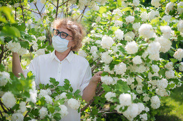 An elderly woman in glasses and a medical mask among the flowers of apple bushes during quarantine....