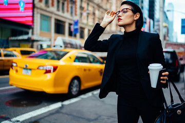 Serious businesswoman in black elegant wear standing on road holding coffee cup looking for taxi, confident female entrepreneur waiting for yellow cab on big city avenue hurrying to get to office.