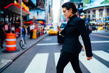 Young successful businesswoman in black suit strolling on crosswalk on Manhattan street, confident female manager holding coffee to go cup with copy space for brand name or label walking on avenue..