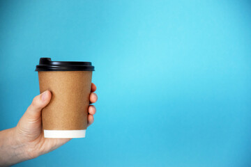 Hand holds cardboard cup of coffee with empty space for text on blue background. Coffee shop or delivery service. Copy space.