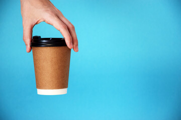 Female hand holds disposable cardboard cup of coffee on blue background. Safe take-out coffee or delivery service. Copy space.