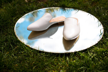 white casual leather slippers on the street, women’s flat shoes comfortable and useful lie on a fashionable round mirror with reflection of shoes, sky and grass