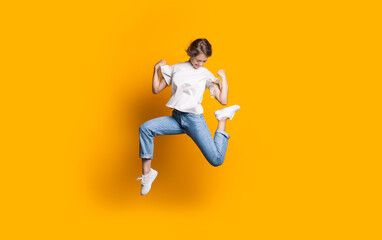 Fototapeta na wymiar Jumping caucasian woman in jeans and white t-shirt smiling and advertising something on a yellow studio wall