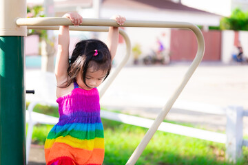 Fototapeta premium Cute Asian little kid girl are wearing colorful clothes hanging on the bars with both hands. Child looked down at the floor. Children diligently exercise with the playground equipment. 3 years old.