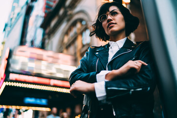 Below view of brunette hipster girl in stylish eyeglasses and trendy wear enjoying spending evening time in New York standing on street with crossed hands.Fashionable female walking in modern downtown