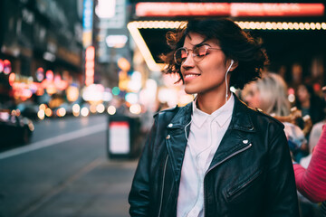 Carefree hipster girl in electronic headphones enjoying Ethno style of music podcast, happy female millennial in earphones listening positive audio playlist and smiling at metropolitan street