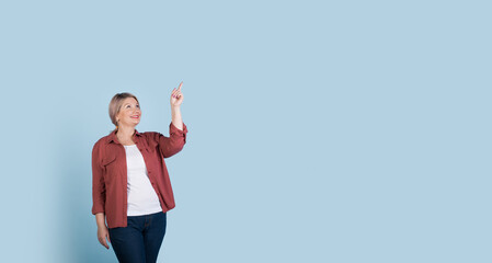 Senior caucasian woman advertising something by smiling and pointing to the blue studio wall with free space