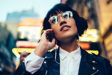 Attractive hipster girl in stylish apparel strolling on urban settings listening to music in earphones, good-looking woman in spending time on city street with night illumination enjoying playlist.