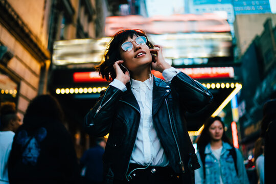 Fototapeta Positive hipster girl in trendy clothes listening favorite song from playlist while strolling on crowded city street in evening, young woman enjoying sound in earphones walking at night downtown