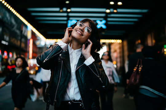 Cheerful beautiful hipster girl in stylish spectacles enjoying favourite music and walk in night city with neon illumination.Happy young woman listening audio song in earphones strolling in New York