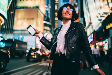 Carefree young woman in eyewear with night city light reflection dancing on crowded street...