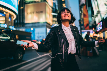 Happy hipster girl in stylish wear spinning on street with night illumination enjoying favorite song in earphones, carefree young woman dancing holding smartphone listening playlist music in downtown.