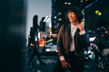 Prosperous party girl dressed in fashionable outfit enjoying nightlife in metropolitan city, happy millennial in optical eyewear and modern headphones listening funny music set during evening walk