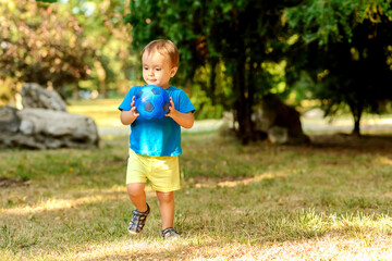 Toddler boy playing alone with blue soccer ball on grass lawn in sunny summer day. Visionary little child holding football ball and thinking about something. Childhood dreams and sports concept