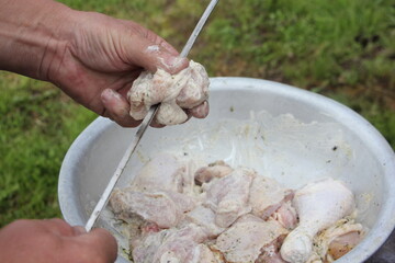 Men hand impales raw chicken meat on a spit from aluminium metal basin, outdoor cooking a poultru shashlik close up on green grass background on summer day