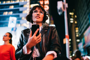 Below view of trendy dressed young woman in stylish eyeglasses strolling on metropolis street with night lights enjoying skyscrapers and listening songs from music playlist in earphones on cellular