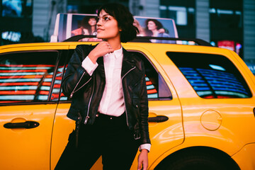 Pondering smiling hipster girl dressed in stylish leather jacket posing on city background with...