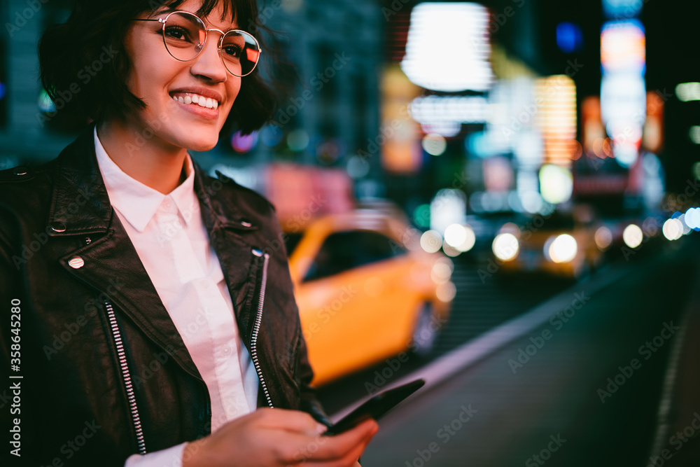 Poster cheerful hipster girl in eyewear using application for calling taxi standing on urban setting with c