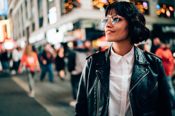Fototapeta premium Carefree hipster tourist in optical spectacles for provide eyes protection enjoying evening walk in New York, attractive Caucasian woman dressed in trendy jacket looking around on Manhattan