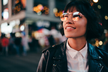 Carefree brunette young woman with short haircut in stylish eyewear for eyes protection fascinated with New York urbanity in Manhattan,youthful female in leather jacket enjoying metropolitan nightlife