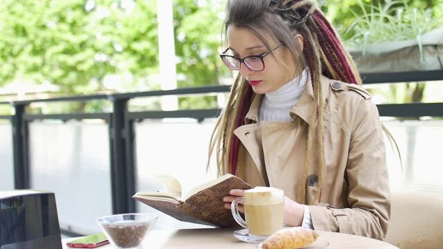 Modern young business woman with long dreadlocks and eyeglasses is writting some notes in her notebook while sitting in a street coffee shop during a day, remote work concept