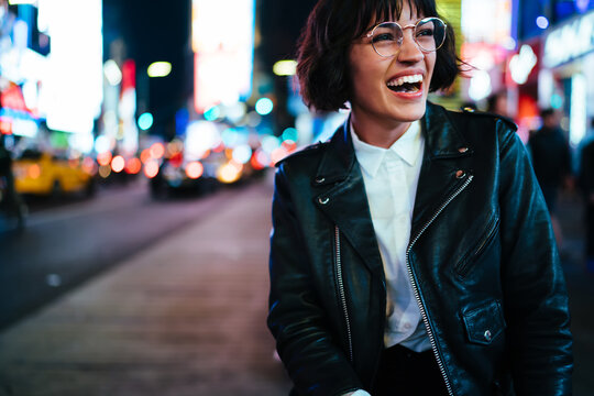 Excited young woman with short haircut laughing at urban setting with night lights enjoying leisure time in New York, happy hipster in stylish wear and eyewear having fun on megapolis street