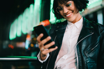 Smiling hipster girl happy about getting message from friend checking mail on phone standing on...
