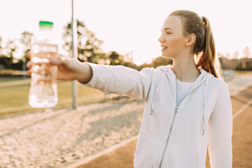athletic female runner standing outdoors at the stadium holding a water bottle. Fitness woman takes a break during a workout