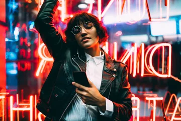 Fotobehang Beautiful brunette woman in trendy apparel and eyewear enjoying nightlife in city listening music in earphones and moving to sound, gorgeous hipster girl dancing outdoors on neon city illumination . © BullRun