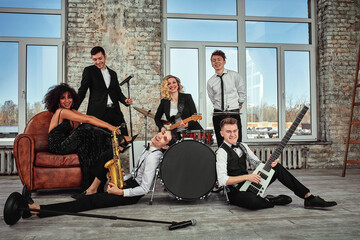 Photo of a lot of ethnic musical group in studio. Musicians and an African American woman soloist posing on camera, during a rehearsal, loft background.