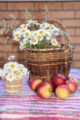 Fototapeta na wymiar A large bouquet of daisies in a wicker basket, a small bouquet - in a pot on a woven striped bright carpet. Ripe juicy apples lie nearby. Vertical photo.