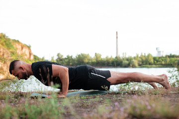 Profile of serene attractive bearded young man in black dress doing stretching sport, yoga, fitness or pilates exercises on green lawn, working out for flexible spine.